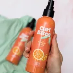 Kézben tartott narancssárga Curl Bites frissítő spray fekete adagolófejjel és narancs mintával,a háttérben elmosódva a Curl Bites Refresh Serum zöld textíliára fektetve.