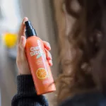 Kézben tartott narancssárga Curl Bites frissítő spray fekete adagolófejjel és narancs mintával, a kép jobb oldalán elmosódva göndör hajú nő sziluettje hátulról.