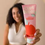 Curl Bites Refreshing Weightless Shampoo sampon rózsaszín tubusban, kézben tartva, göndör hajú nővel a háttérben.