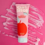Curl Bites Refreshing Weightless Shampoo sampon rózsaszín tubusban, piros alma mintával és kék kupakkal, rózsaszín habos háttér előtt.