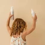 Göndör tincses kislány háttal állva a Nayeel My First habsampon és hajspray flakonjait tartja a magasba bézs háttér előtt.