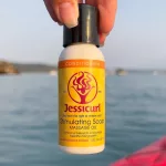 Kézben tartott kis flakonos Jessicurl Stimulating Scalp fejbőrolaj sárga etikettel, tengeri háttér előtt.