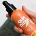Kézben tartott, narancssárga Curl Bites 2 Minute Refresh Serum flakon fekete pumpás adagolóval.