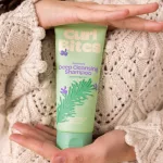 Két kézben tartott világoszöld Curl Bites Rosemary Deep Cleansing Shampoo tubus kötött pulóver előtt.