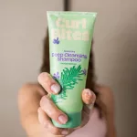 Curl Bites Rosemary mélytisztító sampon világoszöld tubusban, rozmaring mintával habos kézben tartva, a háttérben vizes hajú nő sziluettje látszik.