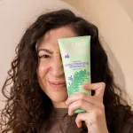 Göndör hajú nő egy világoszöld Curl Bites Rosemary Deep Cleansing Shampoo mélytisztító sampont tart az arca előtt.