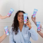 Mosolygó, göndör hajú nő kék köpenyben, aki kék tubusos Curl Bites Deep Conditioner mélykondicionálót tart a kezében.