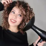 Mosolygó, barna hajú nő a gyűrűs göndör tincseit szárítja fekete Curlsmith diffúzoros hajszárítóval.