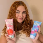 Vörös göndör hajú nő két tubus Curl Bites hajformázó zselét tart a kezében a definiált tincsekért.